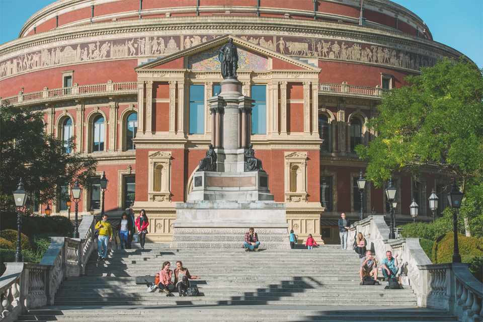 The back entrance of the Royal Albert Hall, with a statue in front of a series of stairs, surrounded by green trees and bushes.